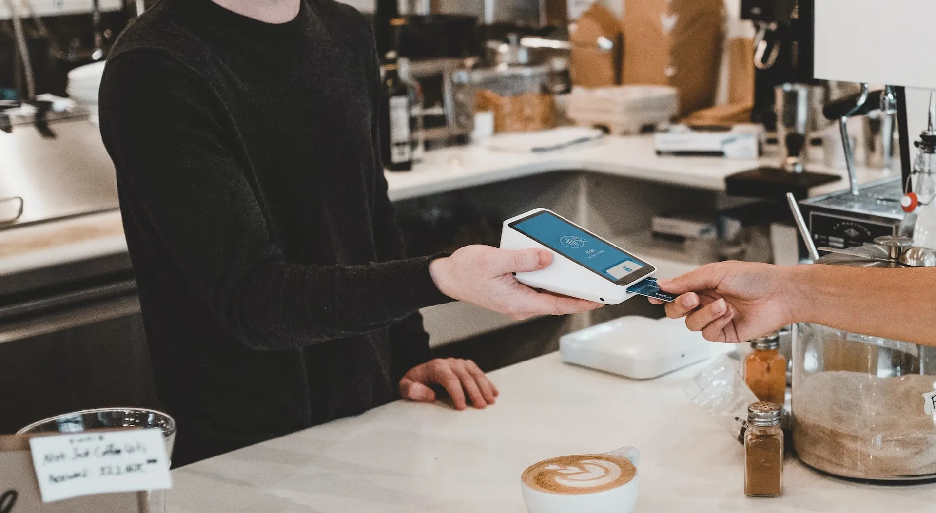 Customer tapping a card on a handheld terminal at a coffee counter